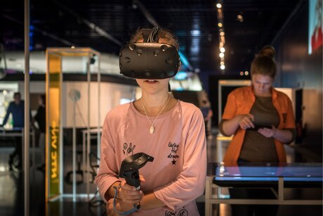 Bild vergr&ouml;&szlig;ern: Person mit Virtual-Reality-Brille und Controller in den H&auml;nden