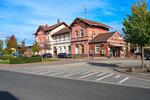 Bild vergr&ouml;&szlig;ern: Ein kleiner l&auml;ndlicher Bahnhof mit rotem Backsteingeb&auml;ude und Cafeteria, umgeben von geparkten Autos, B&auml;umen mit Herbstlaub und einem blauen Himmel. Im Vordergrund ist ein Fu&szlig;g&auml;nger&uuml;berweg.