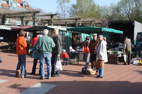 Das Bild zeigt einen belebten Marktplatz im Freien. Mehrere Menschen stehen in Gruppen und unterhalten sich. Im Vordergrund sind zwei Gruppen von Personen zu sehen. Eine Gruppe besteht aus drei &auml;lteren M&auml;nnern und einer Frau, die sich unterhalten. Die Frau tr&auml;gt eine orangefarbene Jacke, w&auml;hrend die M&auml;nner gr&uuml;ne und graue Jacken tragen. Eine andere Gruppe besteht aus zwei M&auml;nnern und einer Frau, die ebenfalls miteinander sprechen. Die Frau h&auml;lt einen Korb in der Hand. 
 
Im Hintergrund sind Marktst&auml;nde mit gr&uuml;nen und wei&szlig;en Markisen zu sehen, an denen verschiedene Waren angeboten werden. Einige Menschen sind dabei, die St&auml;nde zu betrachten oder einzukaufen. Es gibt auch Kinder, die in der N&auml;he der St&auml;nde spielen. Die Umgebung ist von B&auml;umen und Geb&auml;uden umgeben, und das Wetter scheint sonnig zu sein.