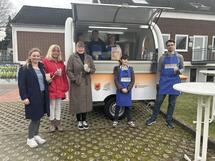 Bild vergr&ouml;&szlig;ern: Eine Gruppe von sieben Personen steht vor einem kleinen Verkaufswagen. Drei Personen sind im Wagen, vier davor. Einige tragen blaue Sch&uuml;rzen mit der Aufschrift 